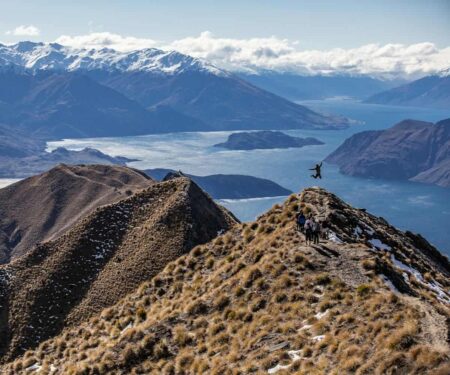 Day Hiking the Southern Lakes Without Packing a Tent A hiker on an exposed Southern Lakes ridgeline overlooking blue alpine lakes and mountain ranges in New Zealand